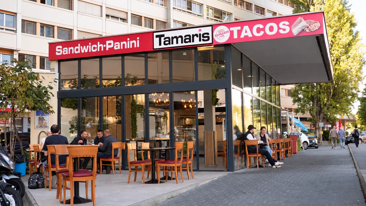 Façade de Tamaris Food à Grand-Lancy : grandes vitres sous l'auvent rouge, enseigne Sandwich-Panini, Tamaris et Tacos, terrasse en bord d'avenue